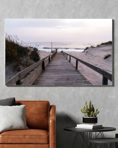A serene beach scene in Portugal. A wooden boardwalk through sand dunes to the ocean, where waves lap the shore under a cloudy sky. Sparse vegetation and evening light create a tranquil atmosphere.