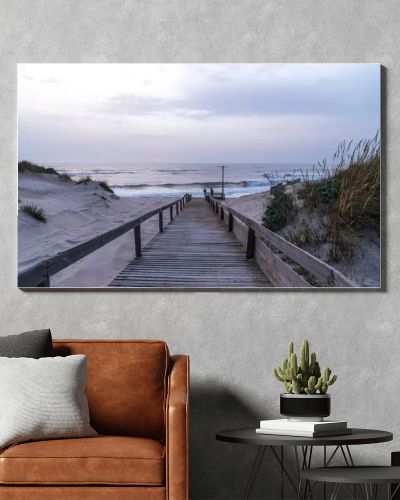 A serene beach scene in Portugal. A wooden boardwalk through sand dunes to the ocean, where waves lap the shore under a cloudy sky. Sparse vegetation and evening light create a tranquil atmosphere.