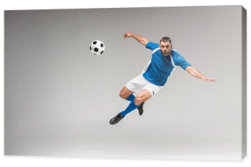 Sportsman looking at camera while jumping near football on grey background
