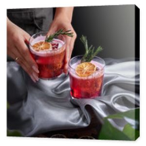 Red drink cocktail of rum and mezcal with lime and cherry juice garnished with dried fruit and green rosemary placed by two female hands