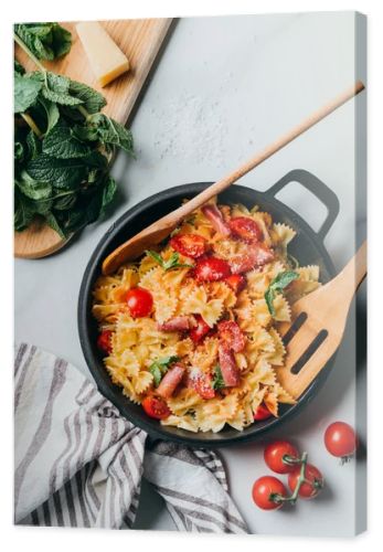 elevated view of pasta with jamon, cherry tomatoes, mint leaves covered by grated parmesan in pan with wooden spatula and spoon on marble table