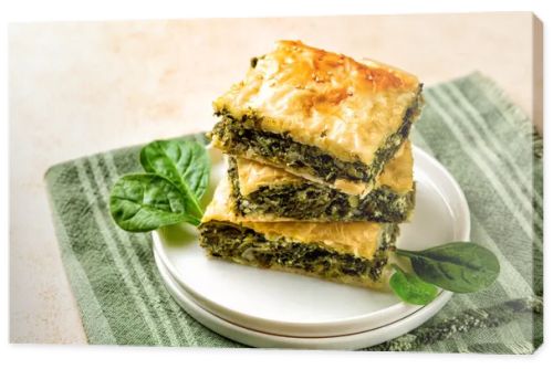 Stack of Spanakopita piece of pie, homemade Greek savory spinach pastry. It contains cheese feta, chopped spinach, green, egg, layered in phyllo or filo dought. Light beige background with green napkin.